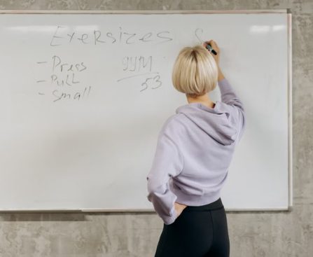 Female writing workout schedule on whiteboard in gym setting.